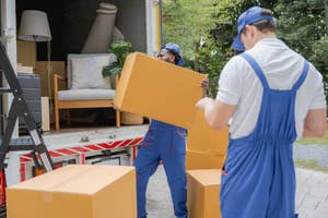 Two movers in uniform unload boxes from a truck. A lamp, chair, and plant are visible in the truck. Their surroundings include greenery