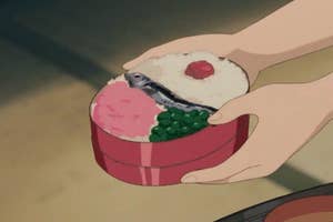 Hands holding a bento box with rice, fish, a red pickled plum, green peas, and a pink side dish. No persons are depicted