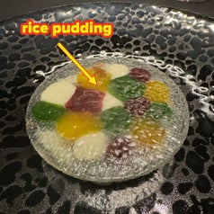 A round gourmet dish features various colorful jelly-like substances arranged on a translucent, lacy surface. A wine glass is seen in the background