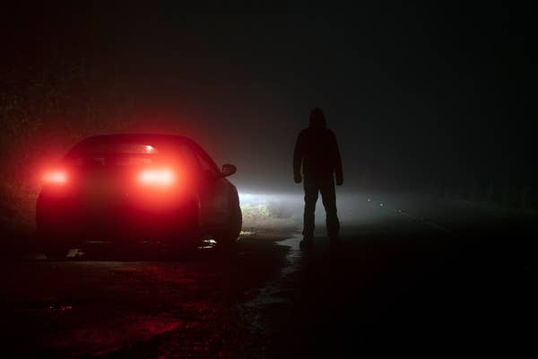A person stands in the dark near a car with rear lights illuminated, facing the car, and is partially visible due to the car's lights