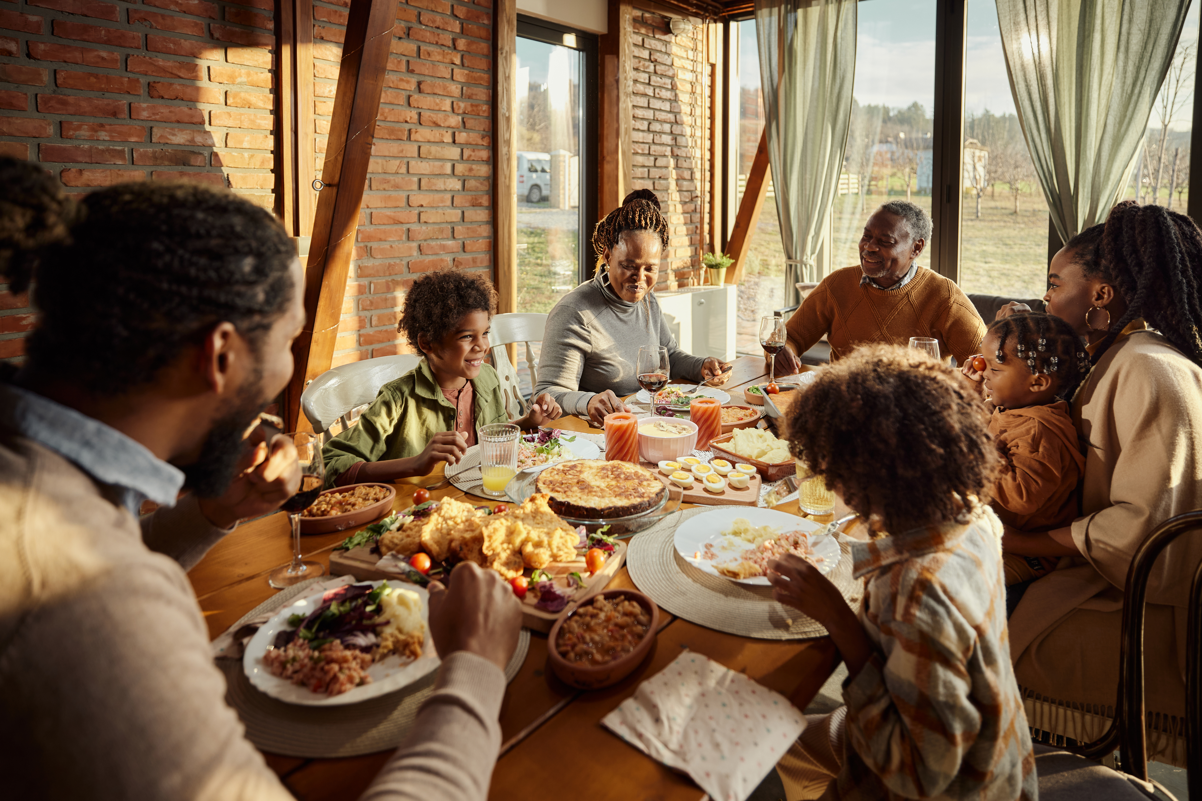 A family of seven enjoys a meal together at a dining table filled with various dishes. The setting is a bright, windowed room