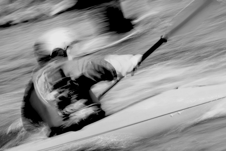 A person in a kayak is navigating through fast-moving water, captured in motion blur. Their face is not visible. No recognizable individuals present