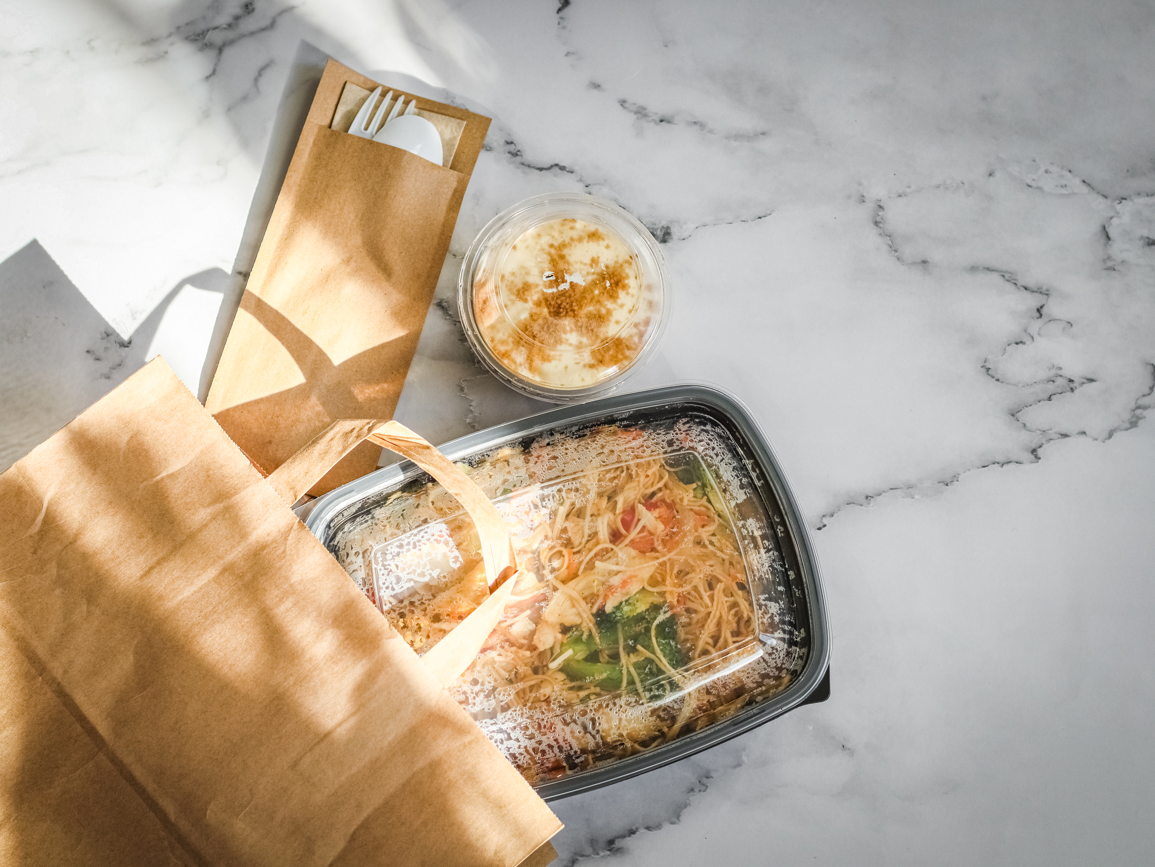 A takeout meal in a black container with a clear lid, a plastic cup with a layered dessert, and a paper bag holding utensils on a marble surface