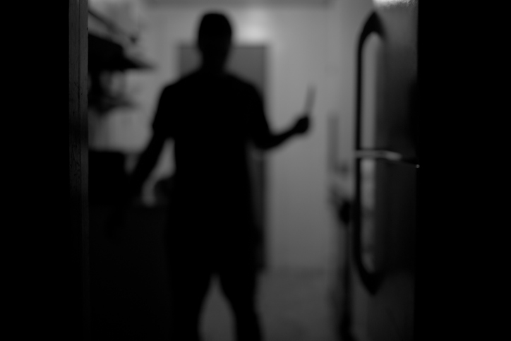 A silhouette of a person holding a knife in what appears to be a dimly lit kitchen. Specific details about the person or surroundings are not clearly visible