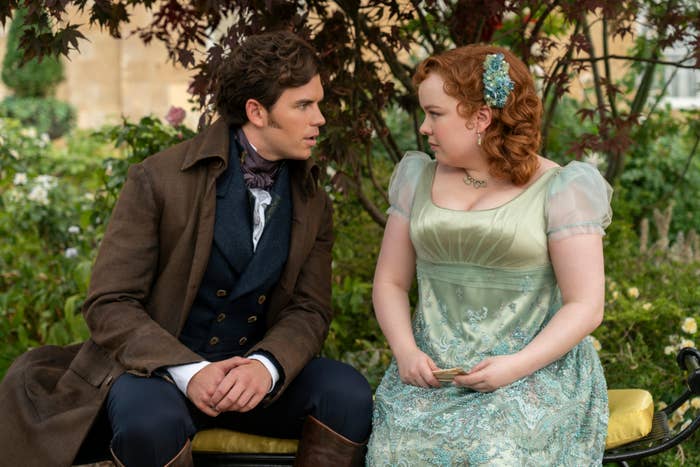 Jonathan Bailey, in a brown coat, and Nicola Coughlan, in an elegant period dress, sit on a bench in a garden from the TV show "Bridgerton"