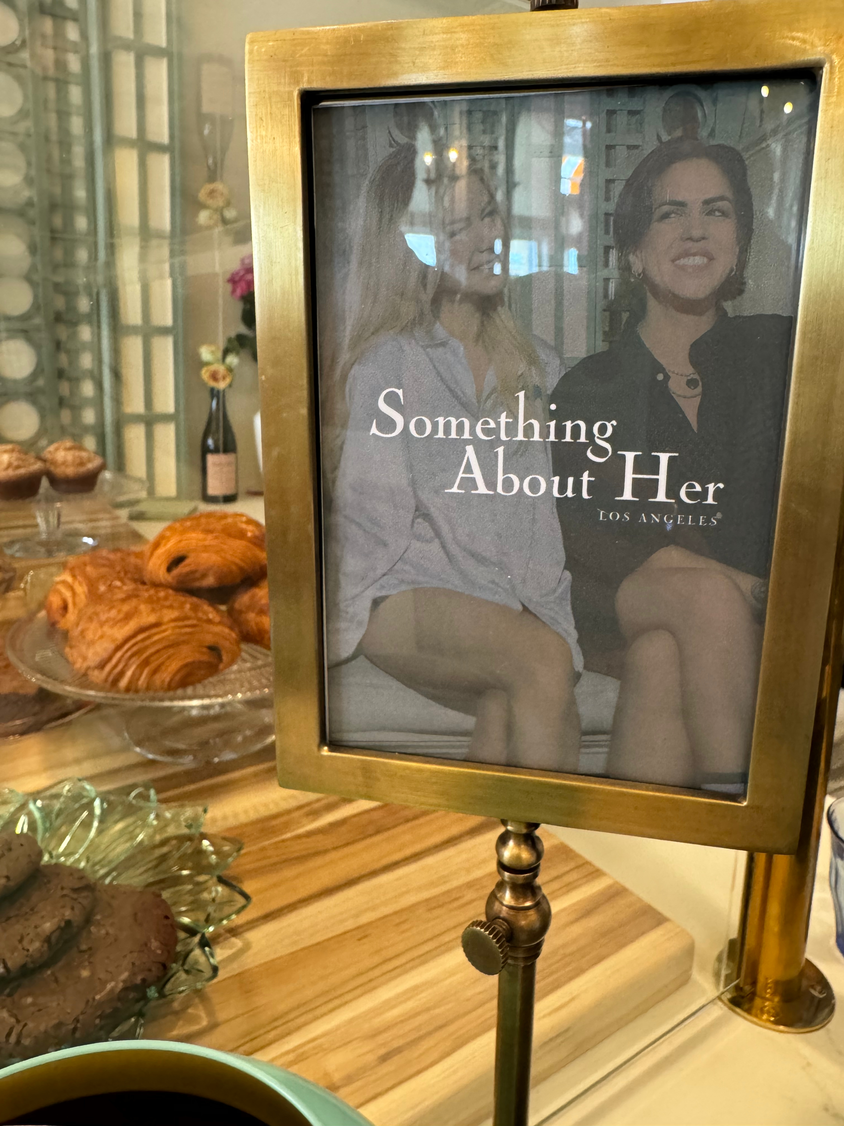 Two women smile in a framed photo with the text &quot;Something About Her Los Angeles&quot; in front of a glass case with pastries