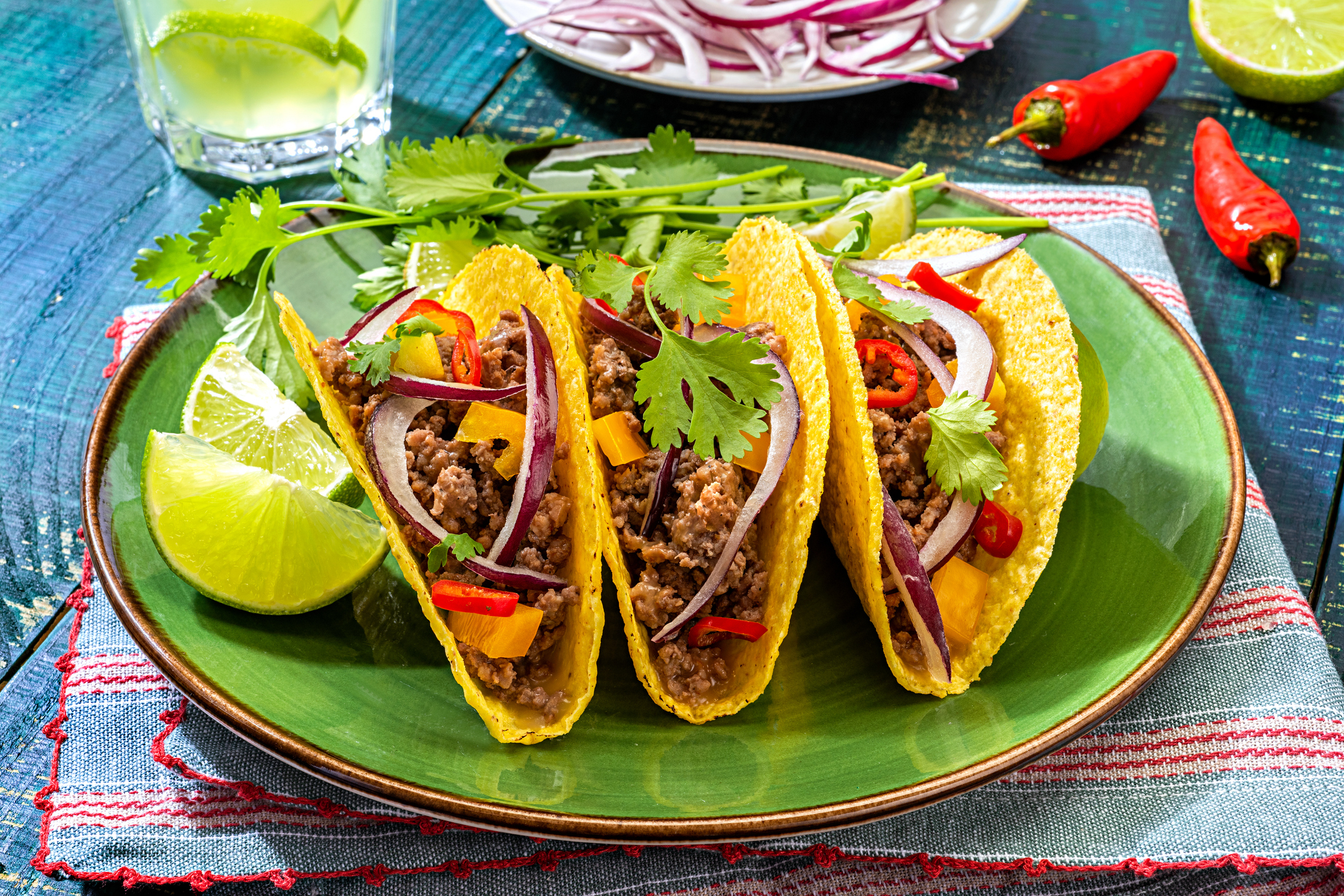 Three tacos filled with ground beef, diced mango, red onions, cilantro, and chili peppers on a green plate with lime wedges, a drink, and a bowl of sliced onions