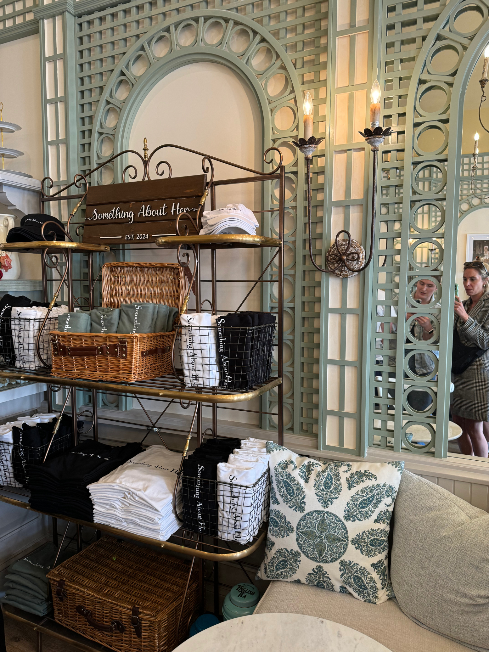 Boutique shop with shelves of folded clothes and a central wicker basket. Two people are visible in the mirror reflection, browsing the store. Sign reads &quot;Something About Her.&quot;