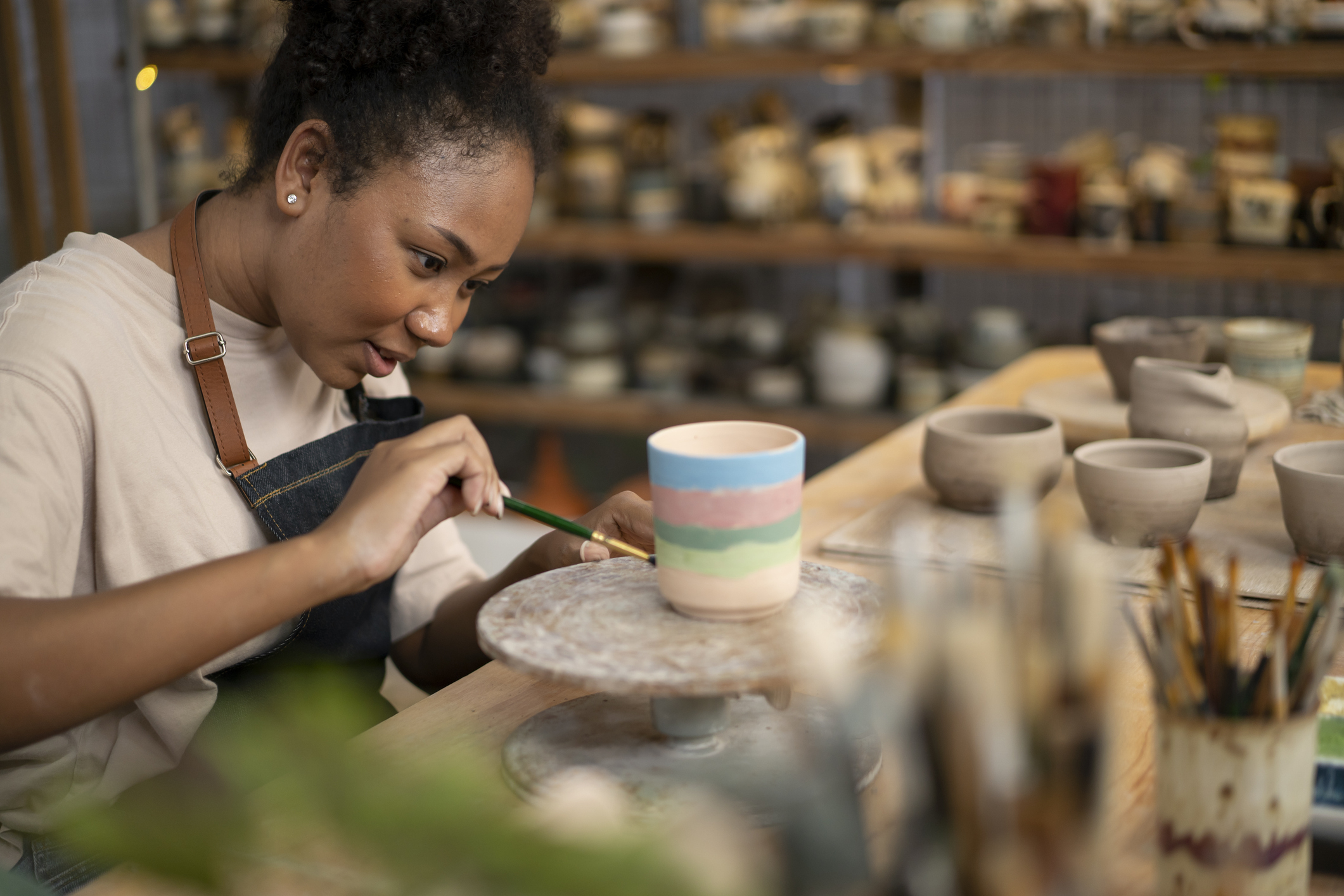 Person painting a ceramic cup with pastel stripes, focused and wearing an apron in a pottery studio with other ceramics on shelves