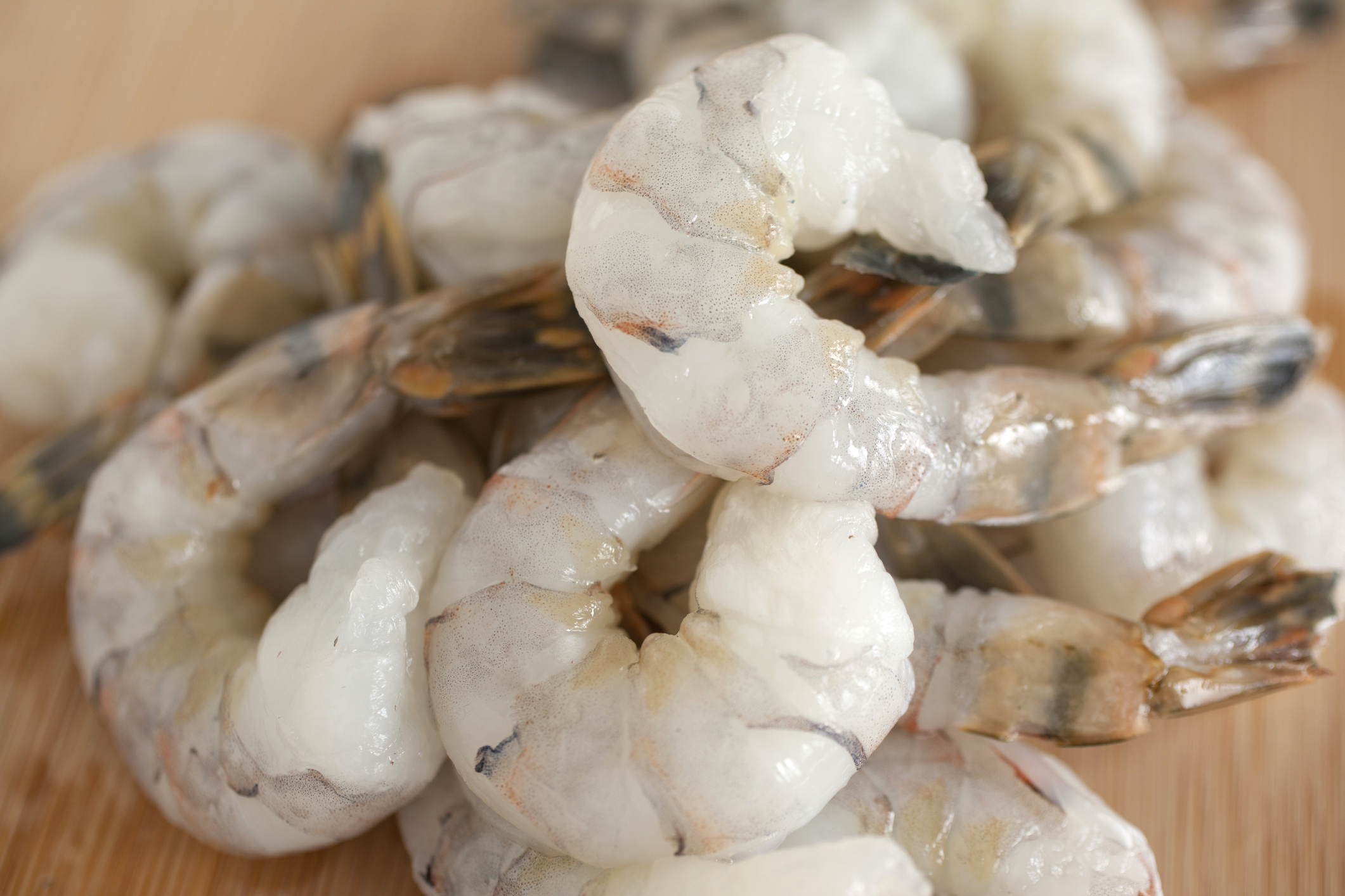 A close-up of raw, peeled shrimp with tails on, piled on a wooden surface