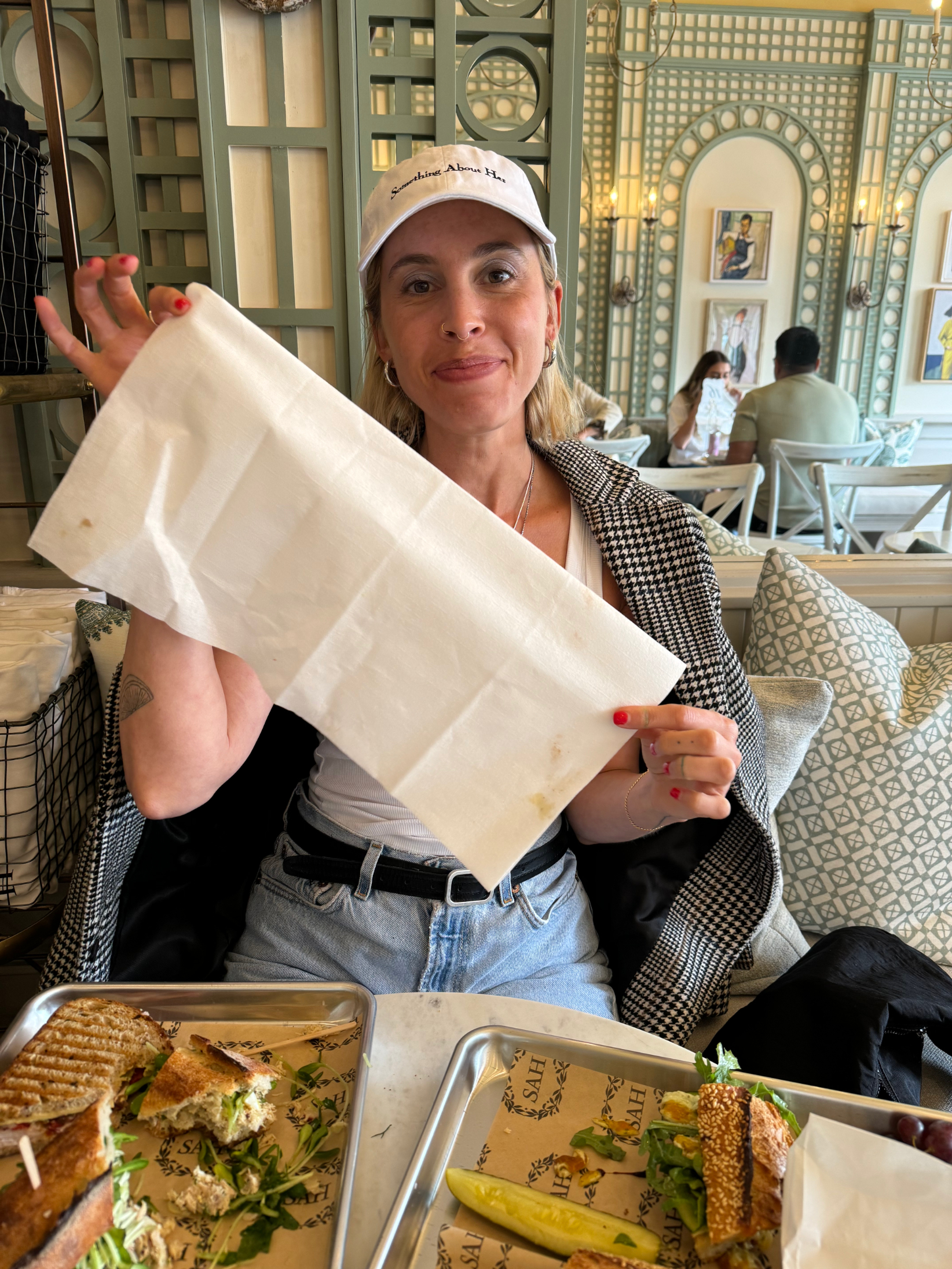 A woman in casual attire and a white cap holds up a napkin while sitting at a table with food. In the background, a man and a child are seated