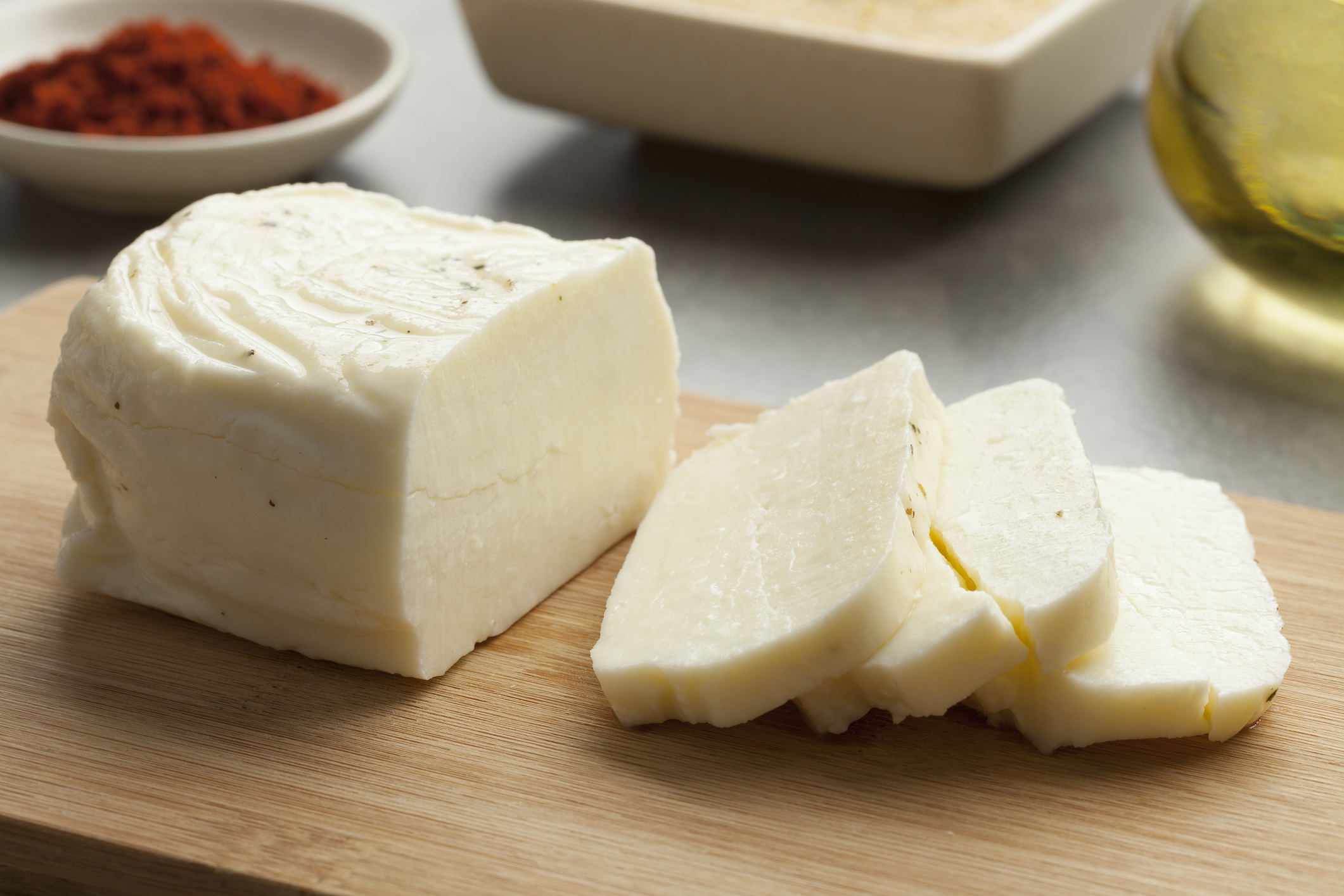 Cheese block with several slices on a wooden board, surrounded by bowls containing spices and oil