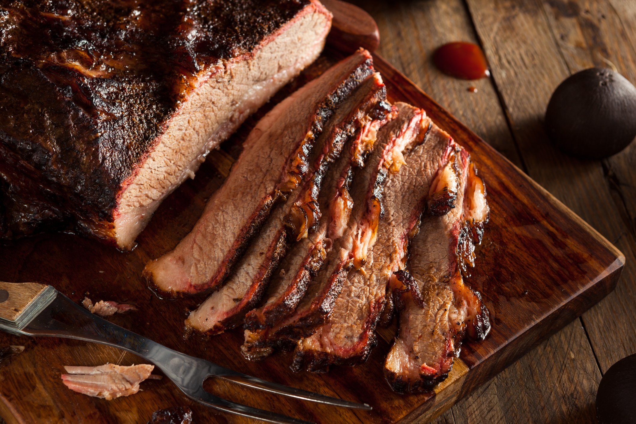 A wooden cutting board with sliced smoked brisket, garnished with a serving fork and a small amount of sauce visible in the background