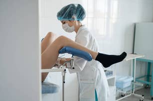 A healthcare professional, wearing a surgical cap and mask, performs a gynecological examination on a patient who is lying on an examination table with her legs in stirrups