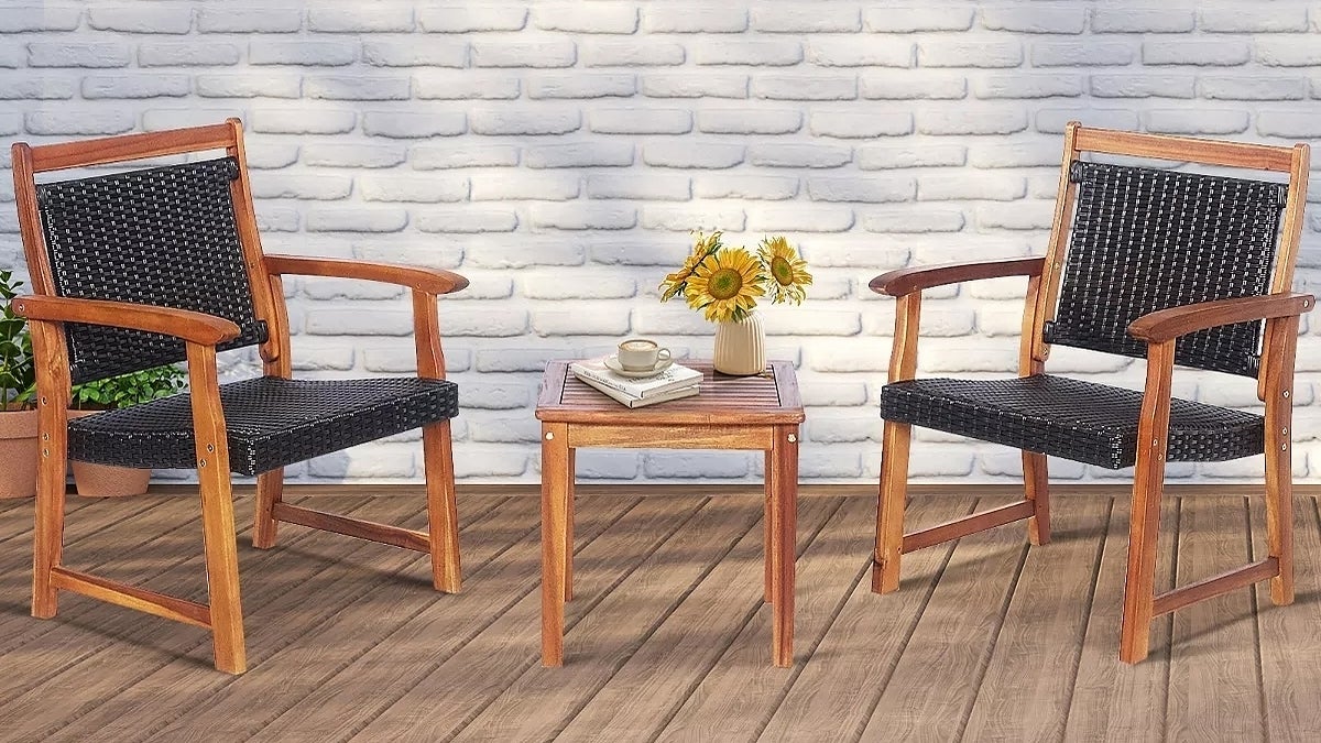 Two wooden patio chairs with dark cushions flank a small matching table holding a vase of sunflowers and a teacup, set against a brick wall with a shuttered window