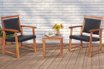 Two wooden patio chairs with dark cushions flank a small matching table holding a vase of sunflowers and a teacup, set against a brick wall with a shuttered window