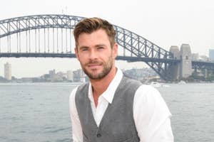 Chris Hemsworth in front of the Sydney Harbour Bridge, wearing a white shirt and a gray vest, smiling at the camera