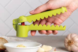 A hand using an alligator-shaped garlic press to crush garlic into a bowl on a kitchen counter with garlic cloves and an onion nearby