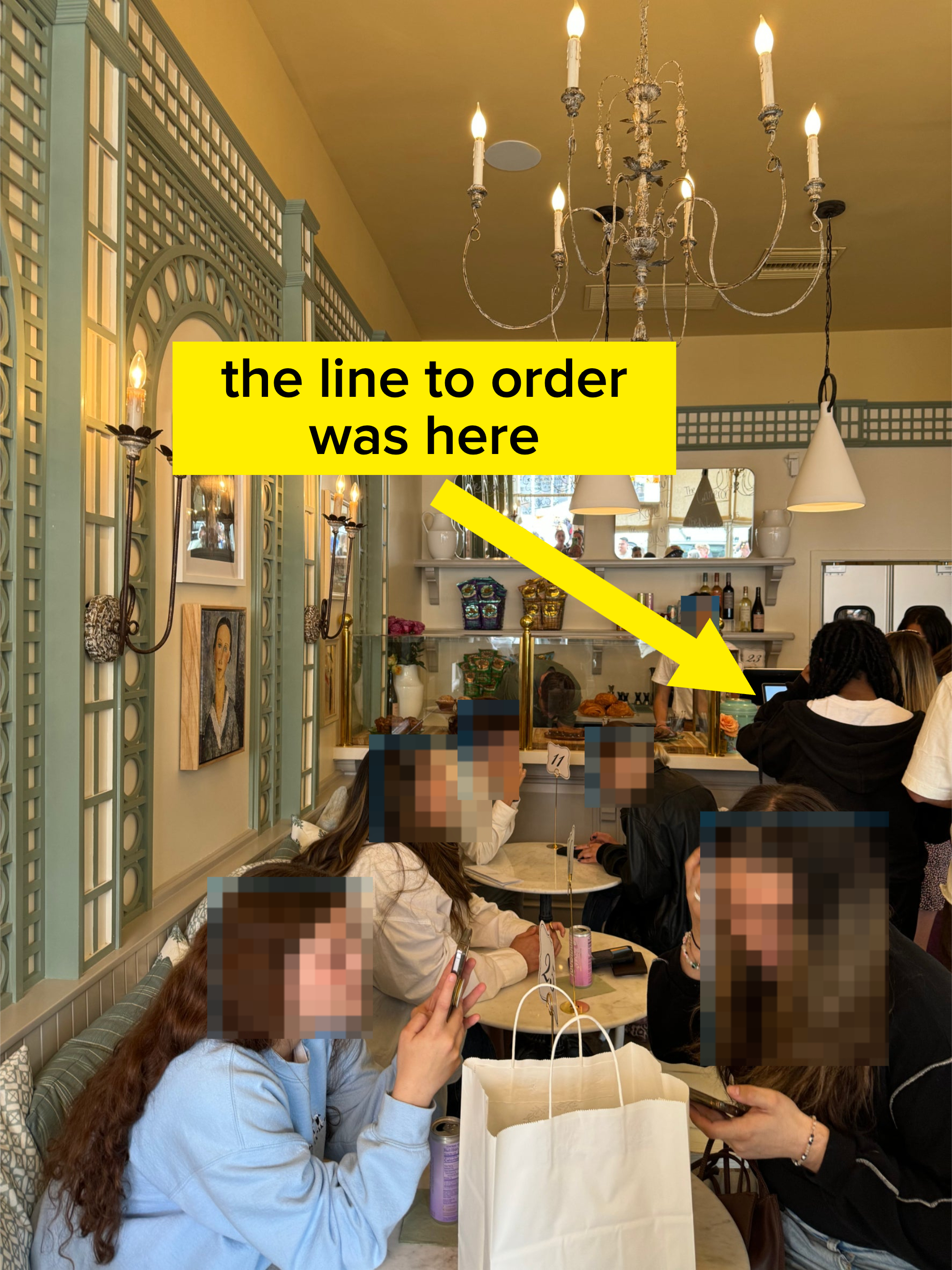 People seated at tables in a cozy cafe, some using phones. A person stands behind the counter taking orders in the background