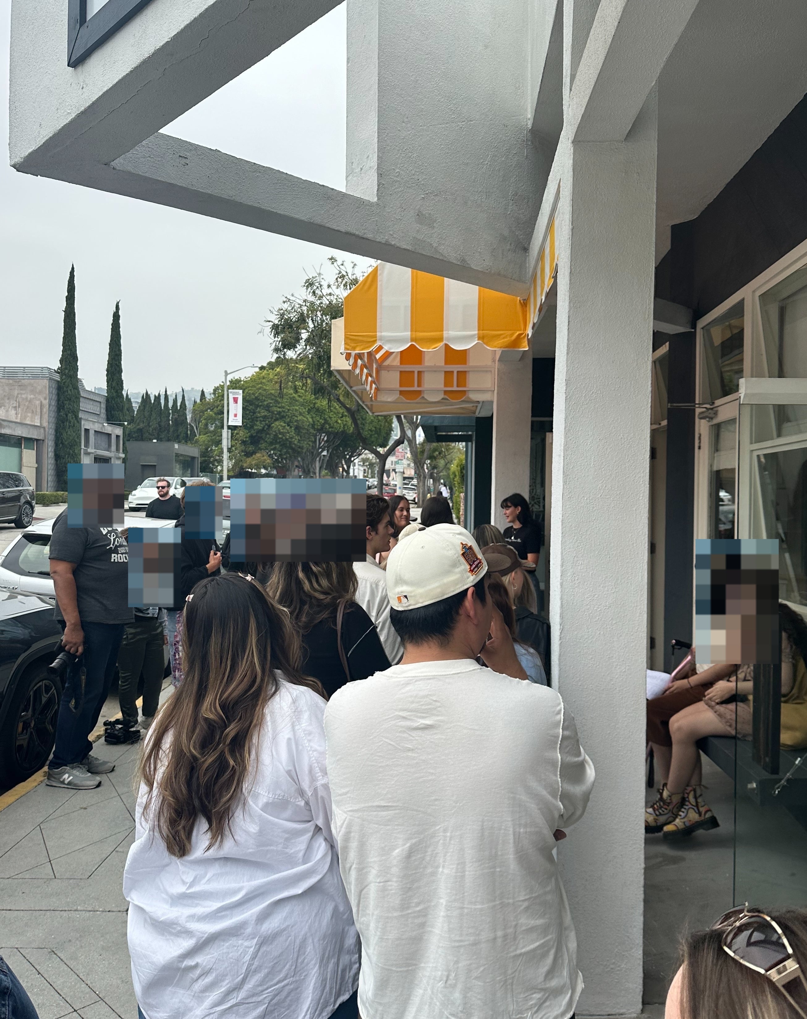People are standing in line outside a store with a yellow-striped awning, waiting to enter. Some are chatting, while others are focused on their phones