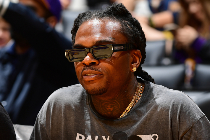 The image shows Gunna wearing sunglasses, a necklace, and a t-shirt, sitting at an event