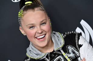 JoJo Siwa smiling, wearing a studded jacket with sequin details, a grey hoodie, and a high ponytail with decorative studs