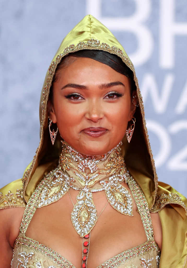 Joy is on the red carpet wearing a bejeweled, hooded outfit with ornate detailing. She is smiling