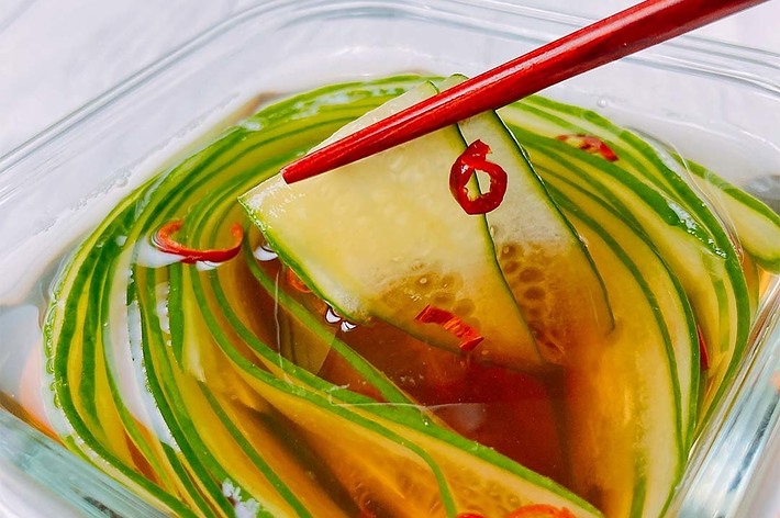 ガラスの器に盛られたスライスしたキュウリの酢漬け。赤い箸で一片を持ち上げている。