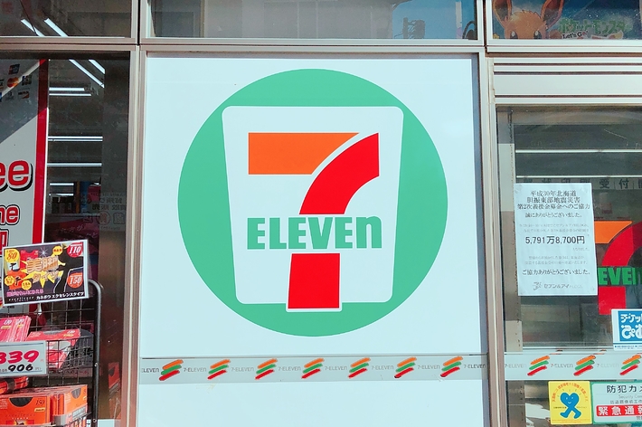 セブンイレブンのロゴが入った看板が店舗の窓に掲示されています。周囲には様々な店舗の広告が見えます。
