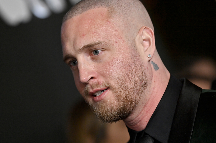 Chet Hanks in a close-up shot, wearing a dark suit at a public event, sporting a shaved head and a neck tattoo