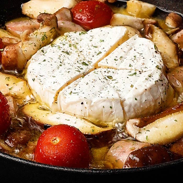 フライパンの中で、丸いカマンベールチーズとトマト、様々なキノコが調理されている様子。