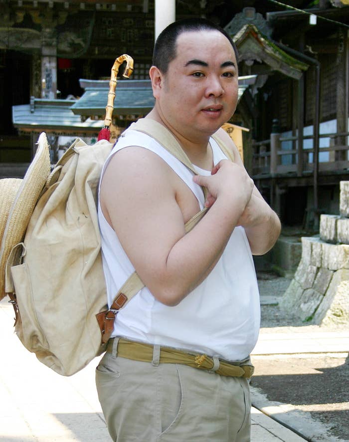 男性がタンクトップとベージュのズボンを着用してバッグを背負い、神社の前に立っている。