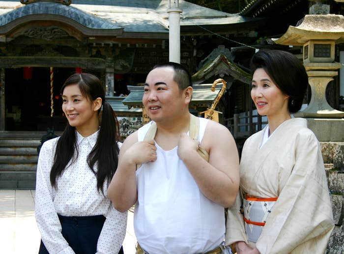 中央に白い服を着た男性が、左側の白いシャツの女性と右側の和装の女性とともに、日本の伝統的な建物の前で微笑んでいます。