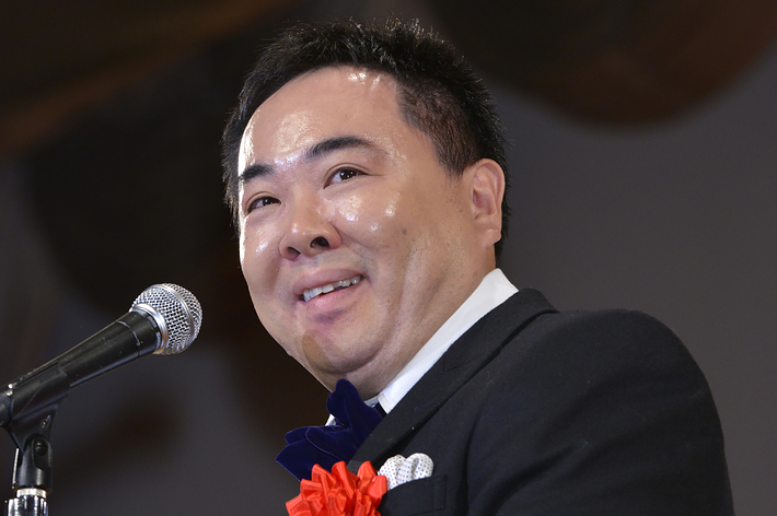 男性がタキシードを着て、マイクの前で笑顔で話をしている。胸に赤いリボンの名札が付いている。