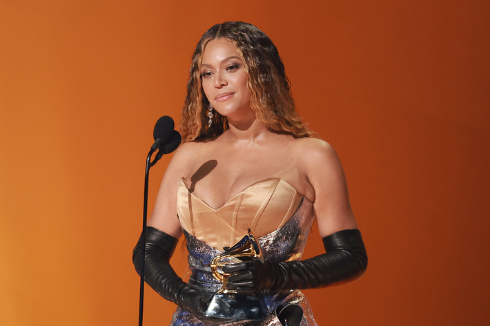 Beyoncé, dressed in an off-the-shoulder gown with long gloves, holding a Grammy award while speaking at the microphone