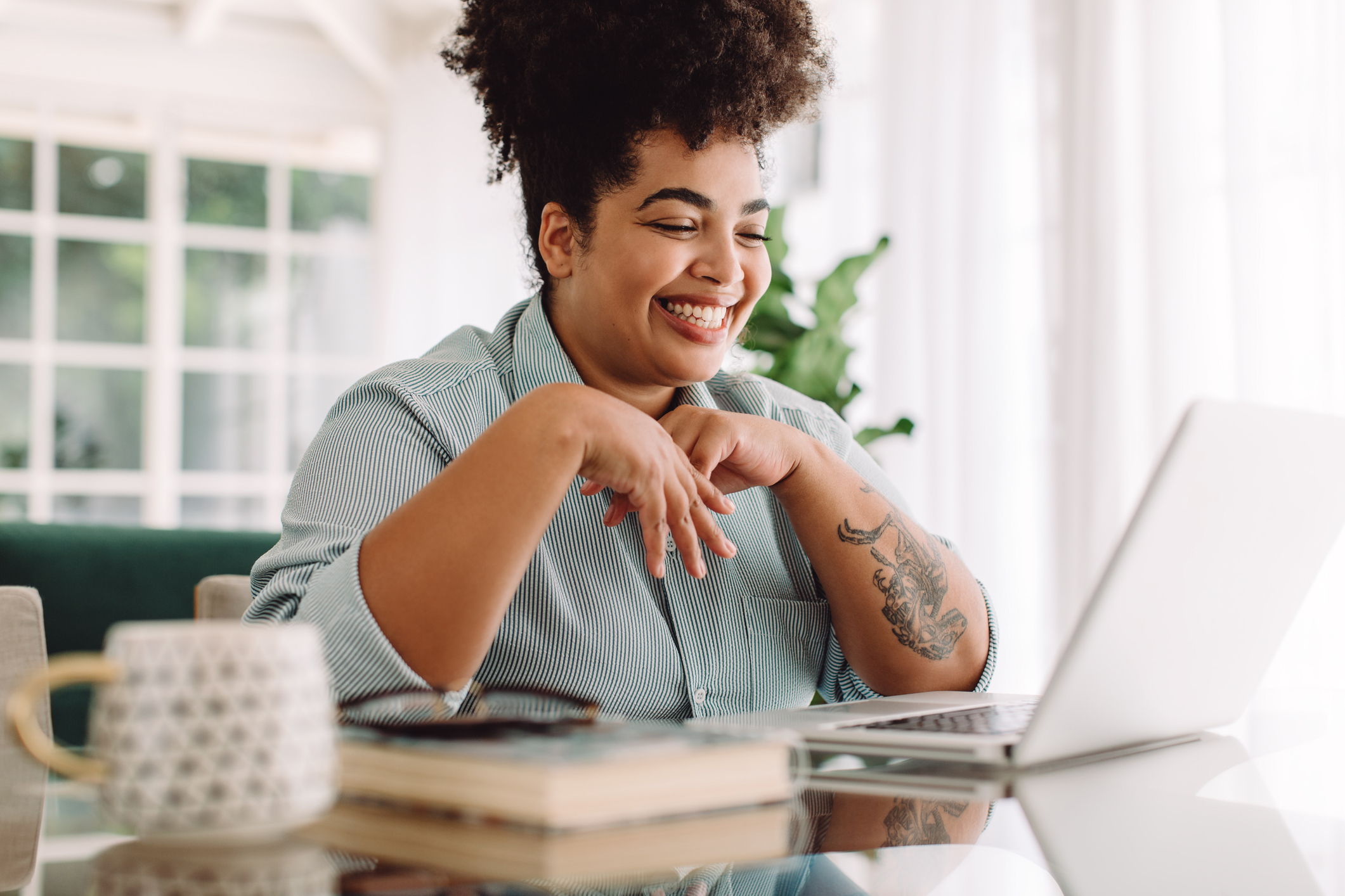 Smiling person with curly hair and tattoos on their arms, wearing a striped shirt, typing on a laptop at a desk with books and a mug