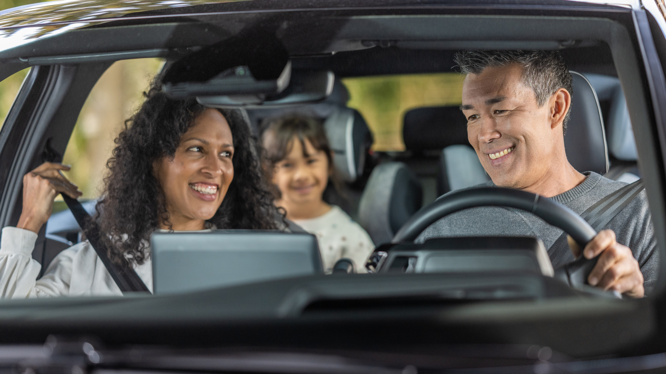 A man and a woman smile while sitting in a car, with a child smiling in the back seat. Everyone is secured with seat belts, and they seem happy and relaxed