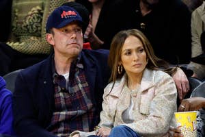 Ben Affleck, Jennifer Lopez, and child sit courtside at a basketball game, with Ben wearing a baseball cap and flannel shirt, and Jennifer in a stylish jacket