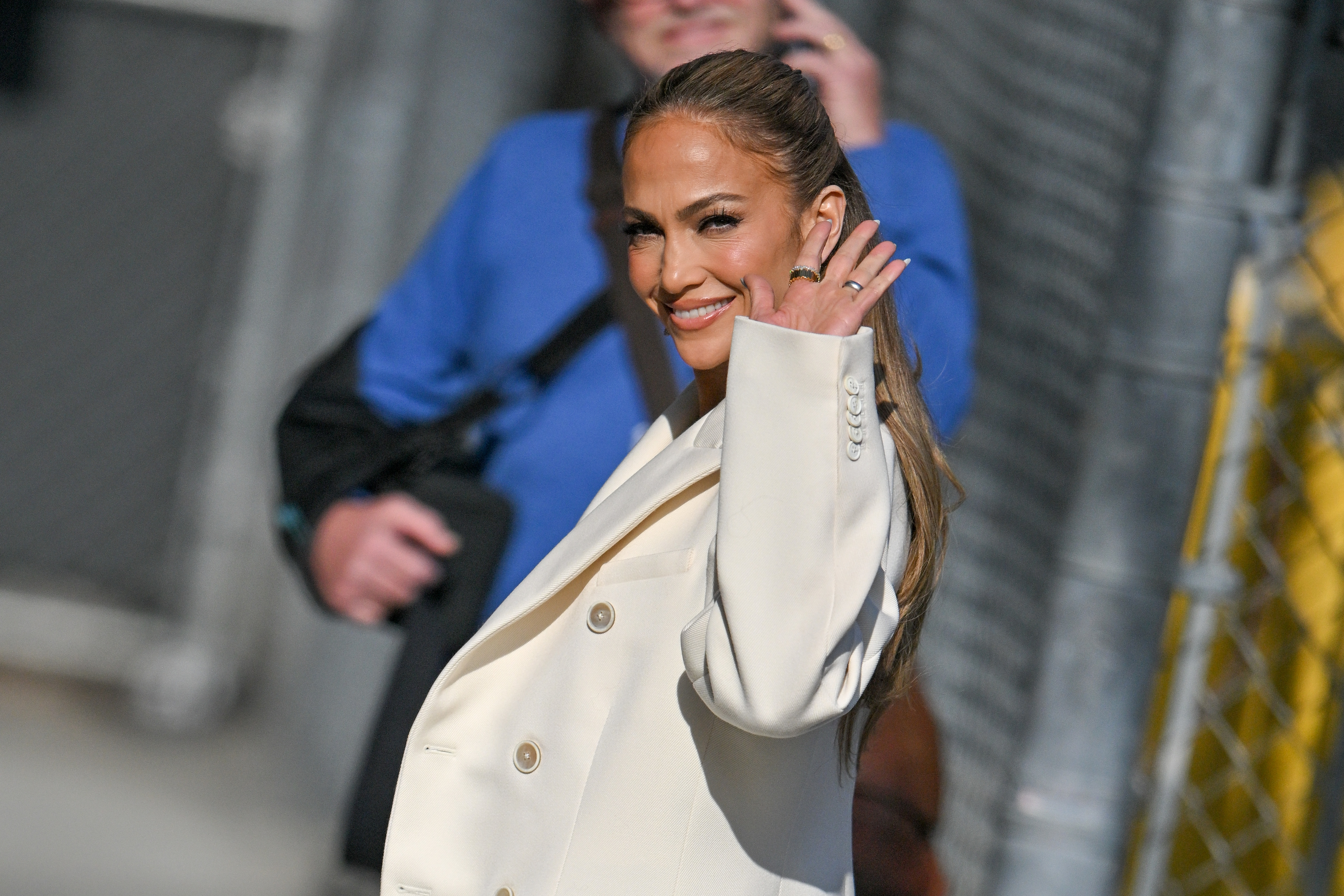 Jennifer Lopez wearing a stylish white blazer, smiling and waving while walking outside