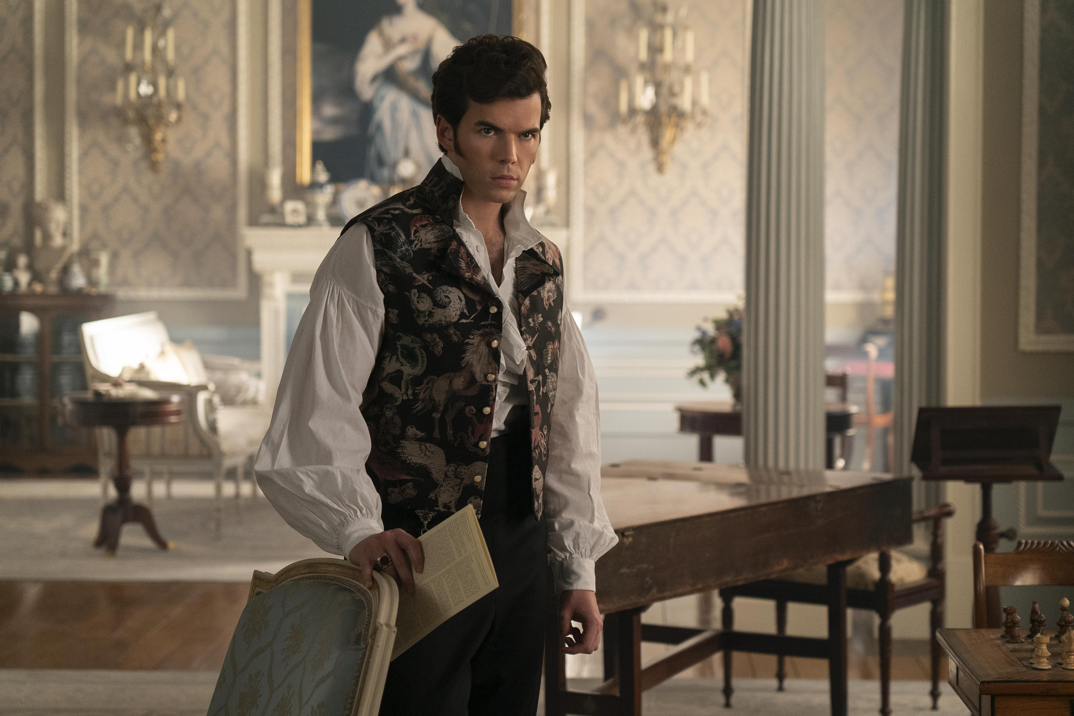 Colin in a period costume with a patterned vest and collared shirt, holding a chair and a document in a lavish, historic room setting