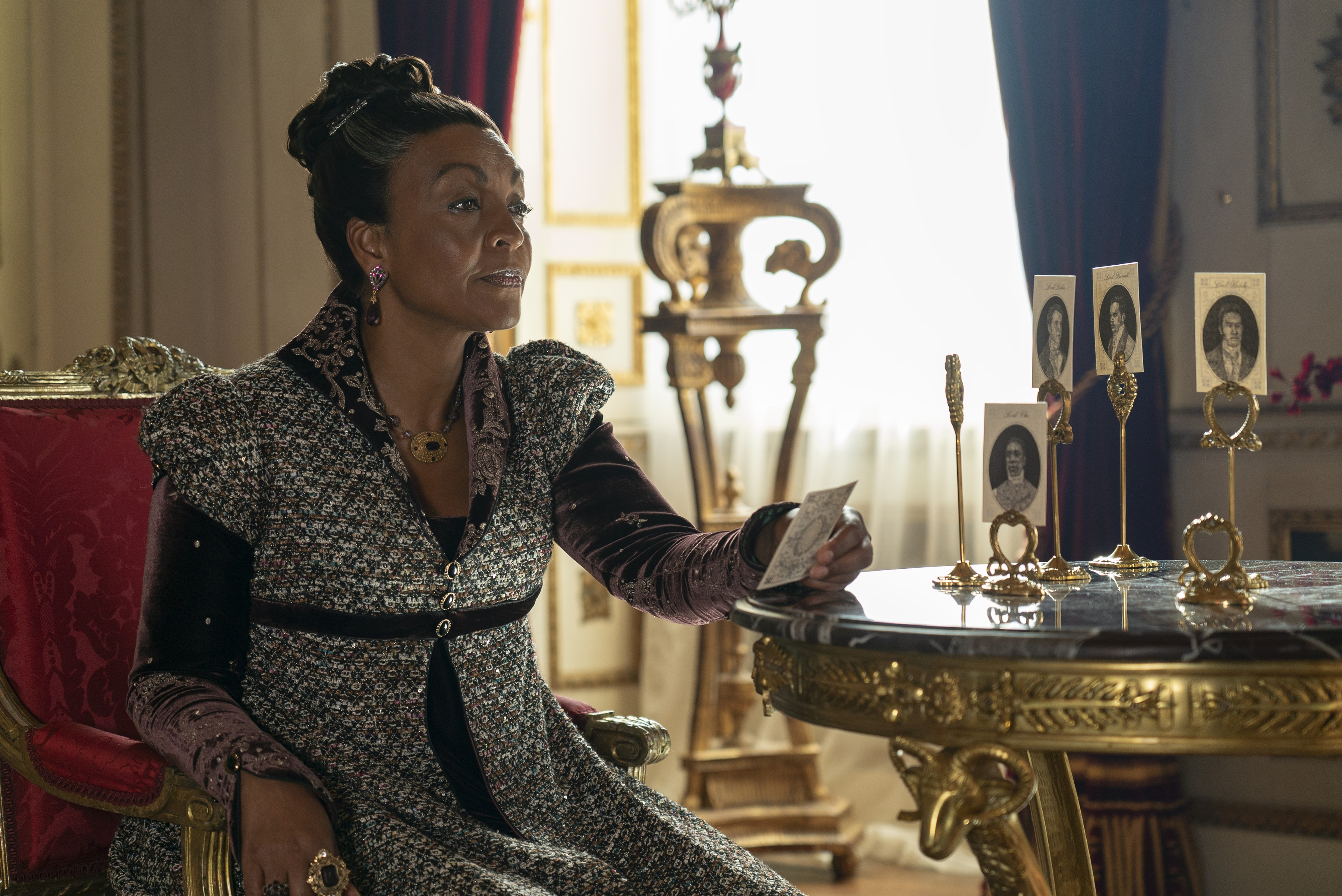 Adjoa Andoh dressed in a Regency-style outfit, seated on a chair, holding a card, with photos on a table in a regal room