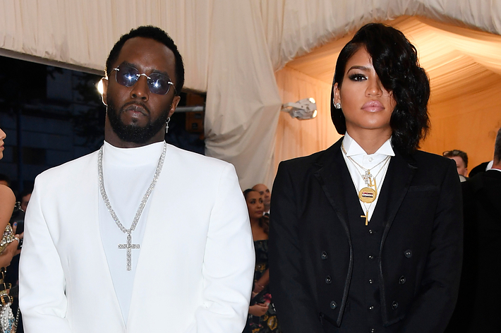 Sean Combs in a white suit and Cassie Ventura in a black suit attend a formal event with other attendees in the background