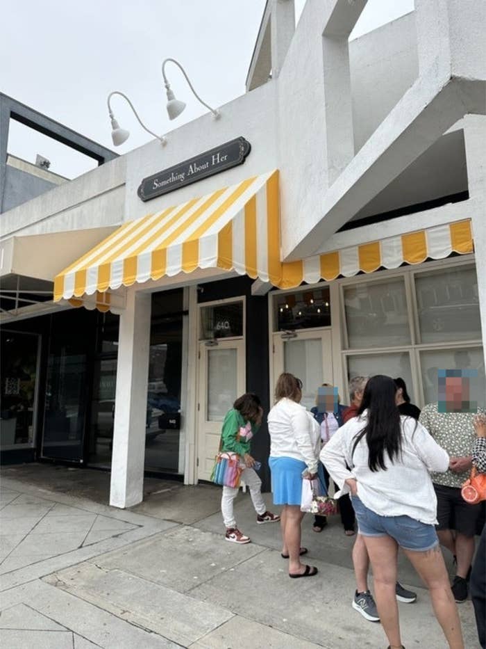 People standing outside called &quot;Something About Her.&quot; Some are talking and others are looking at their phones