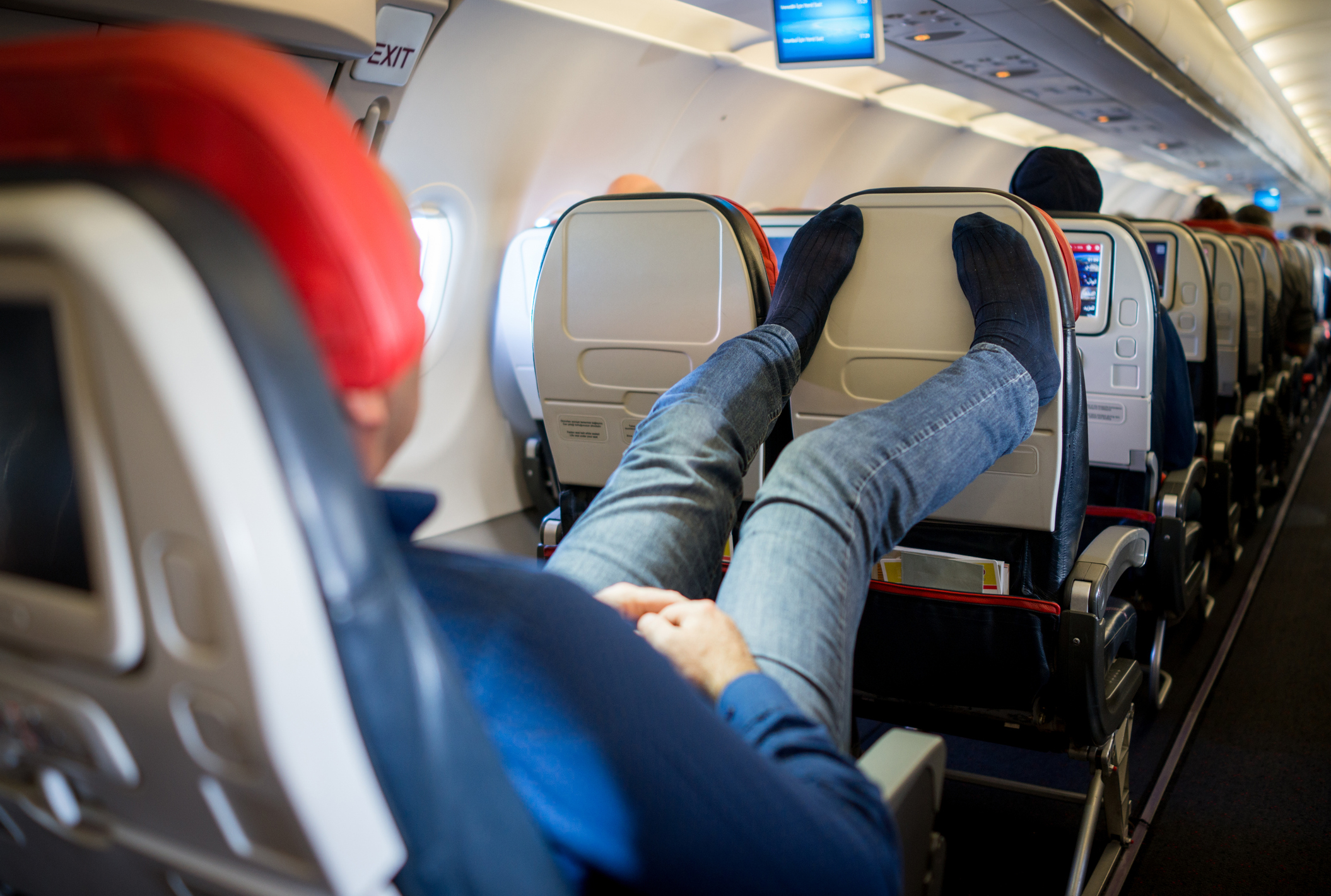 Person reclining in an airplane seat with legs extended over the empty seats in front, showing their sock-covered feet. Seats and screens are visible