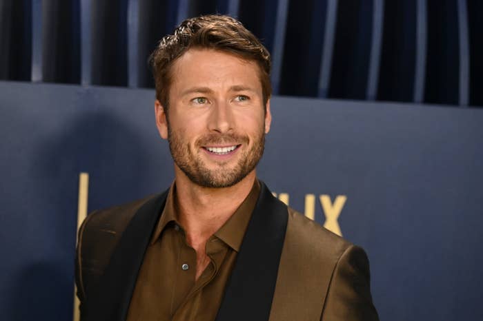 Glen Powell, wearing a brown suit with a black lapel, smiles while standing against a dark background at a celebrity event