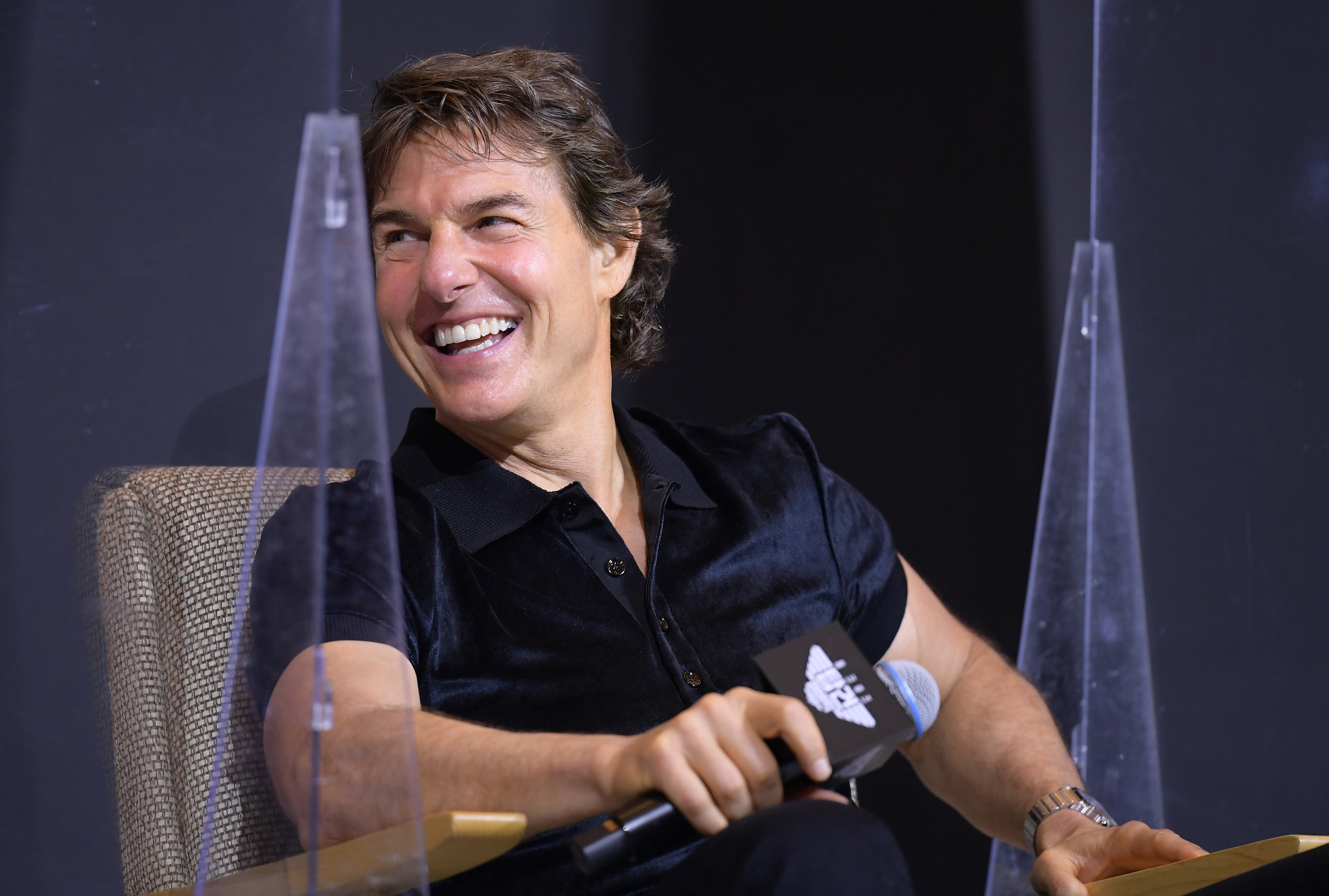 Tom Cruise, seated and laughing, holds a microphone while attending an event. He is wearing a short-sleeved, button-up shirt
