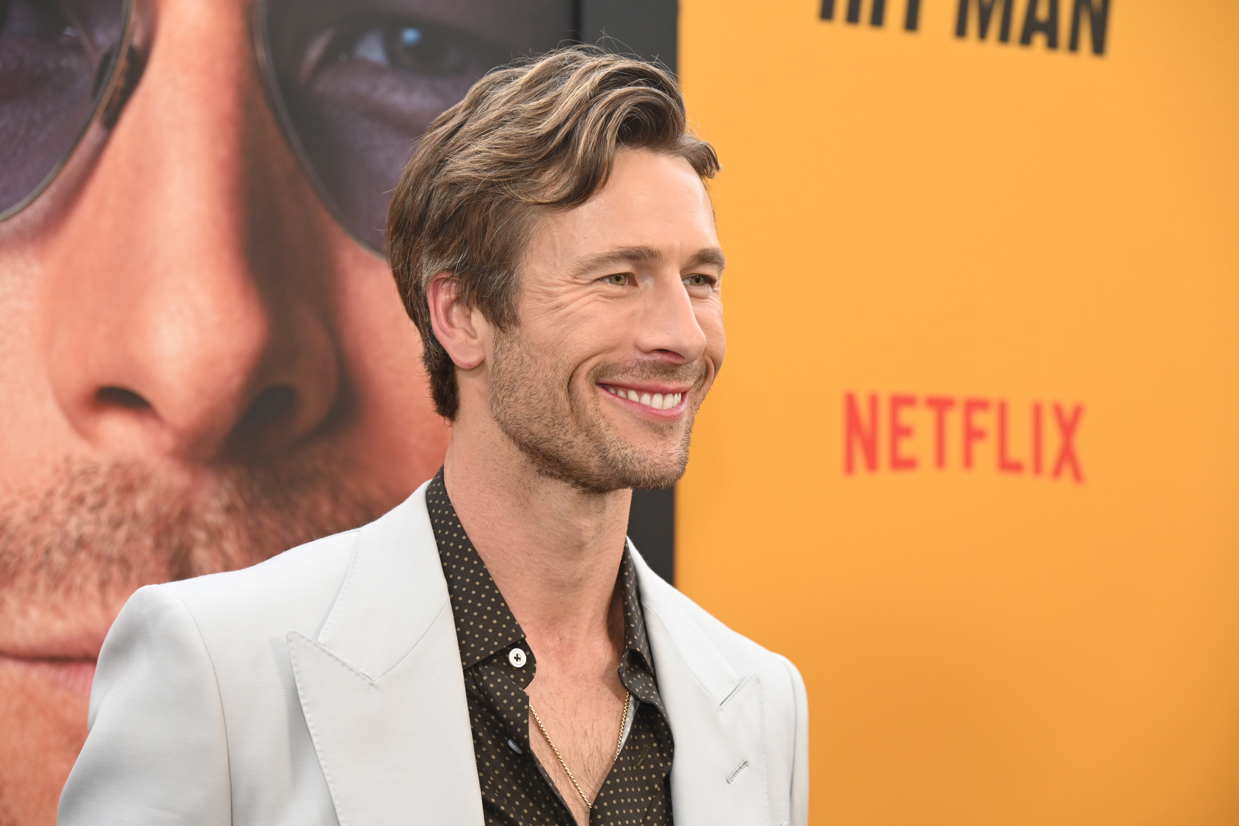 Glen Powell smiles on the red carpet, dressed in a light-colored blazer over a button-down shirt with a dotted pattern, with a poster and Netflix logo in the background