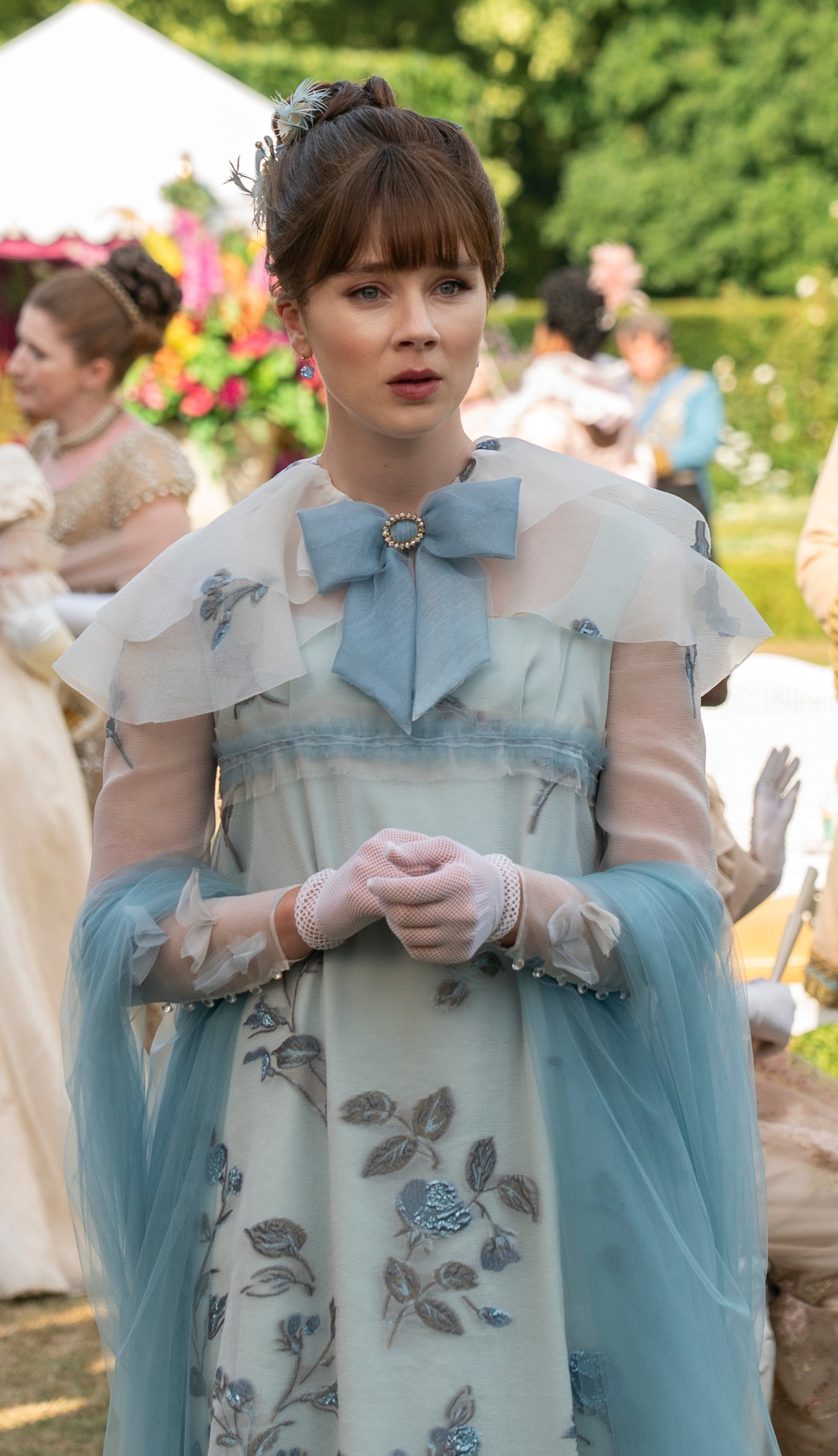 Period-dressed Eloise stands in the foreground at an outdoor gathering, wearing dress with layers of sheer fabric, a shawl, a bow, and embroidered leaves