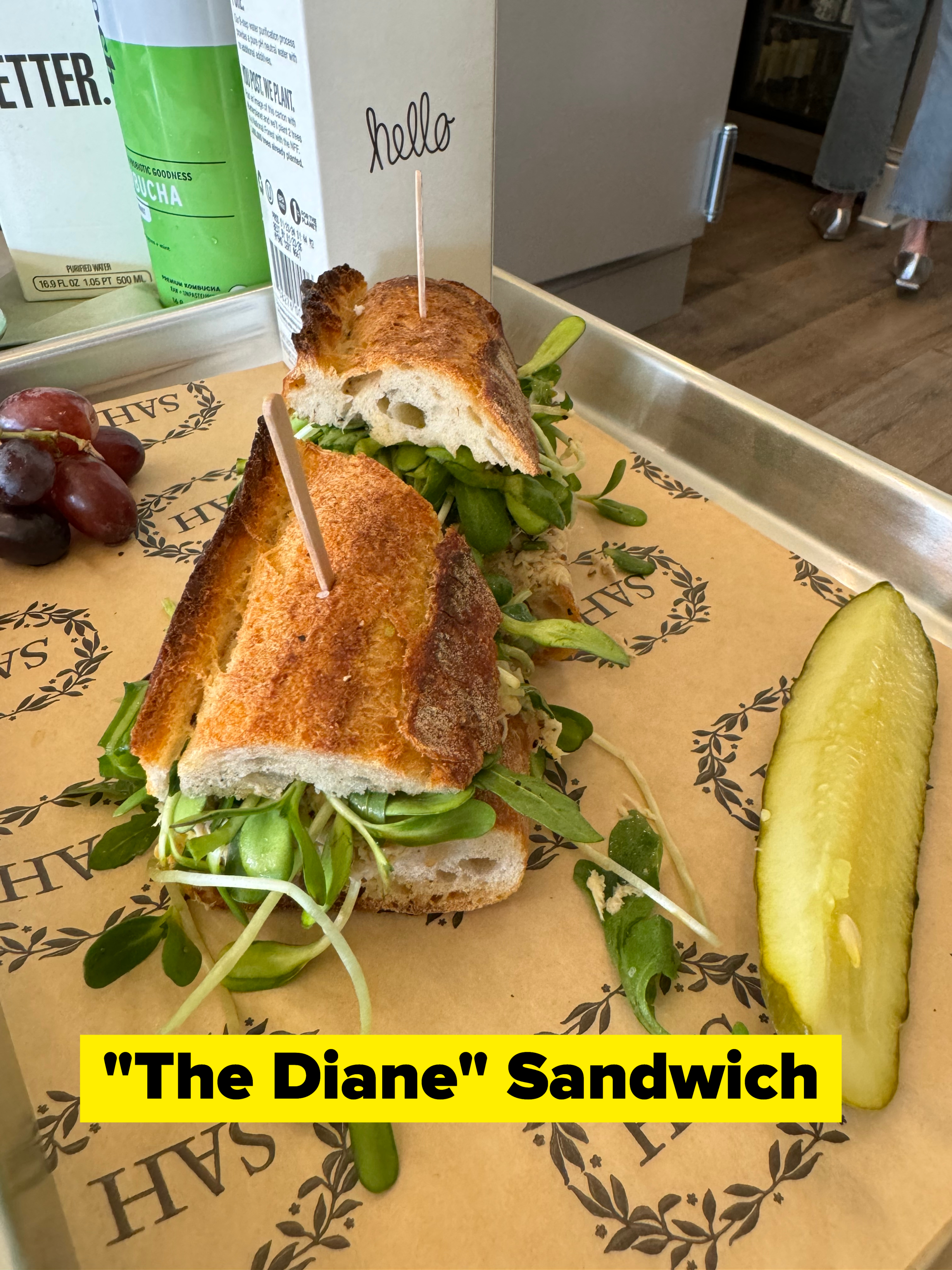 A freshly made baguette sandwich with greens and a pickle slice on a tray. Grapes and a carton of matcha are also visible. The sandwich has a toothpick with a &quot;hello&quot; sign