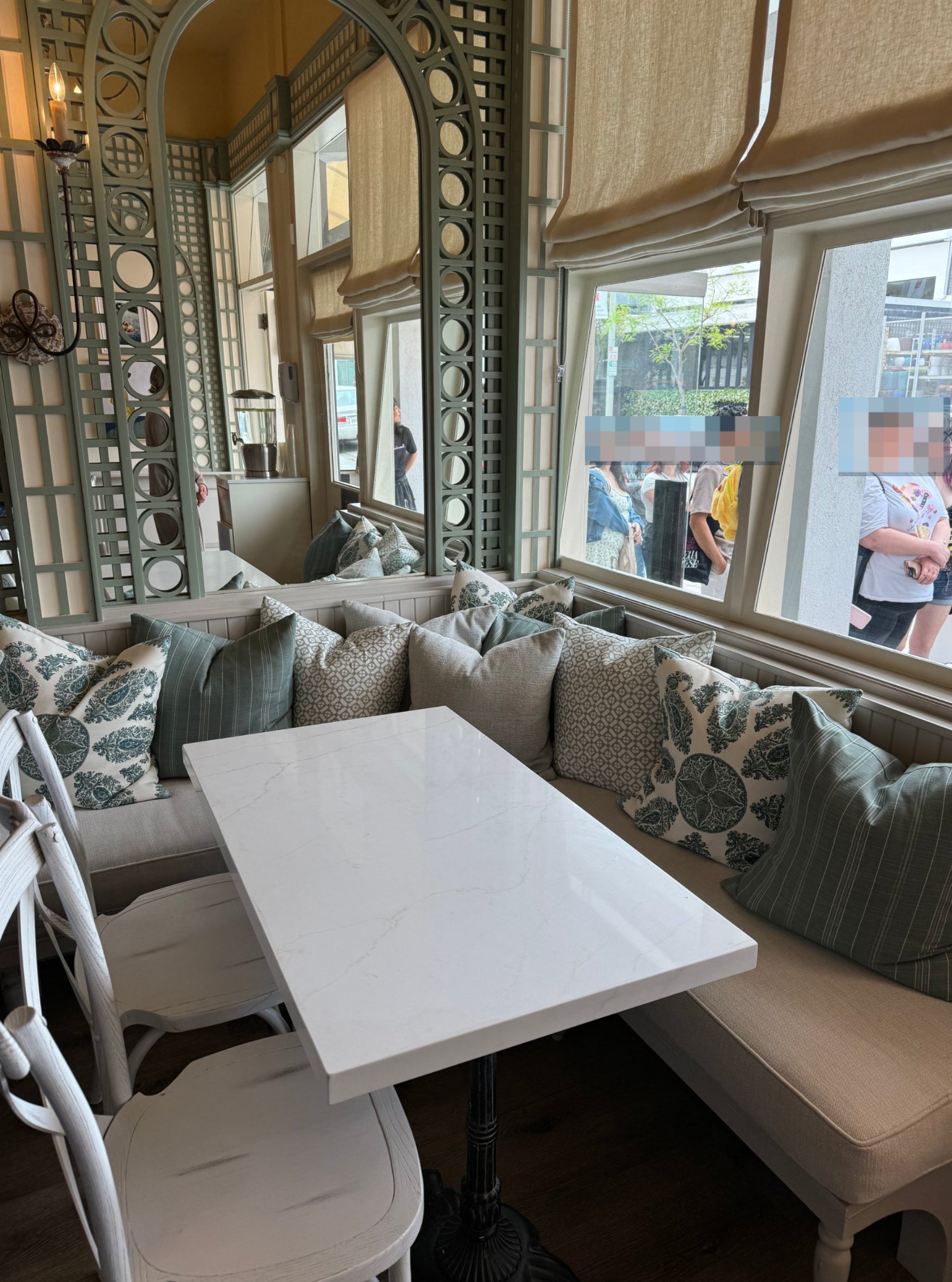 A cozy cafe corner with a white marble table, cushioned bench seating, decorative pillows, and large windows showing people standing outside